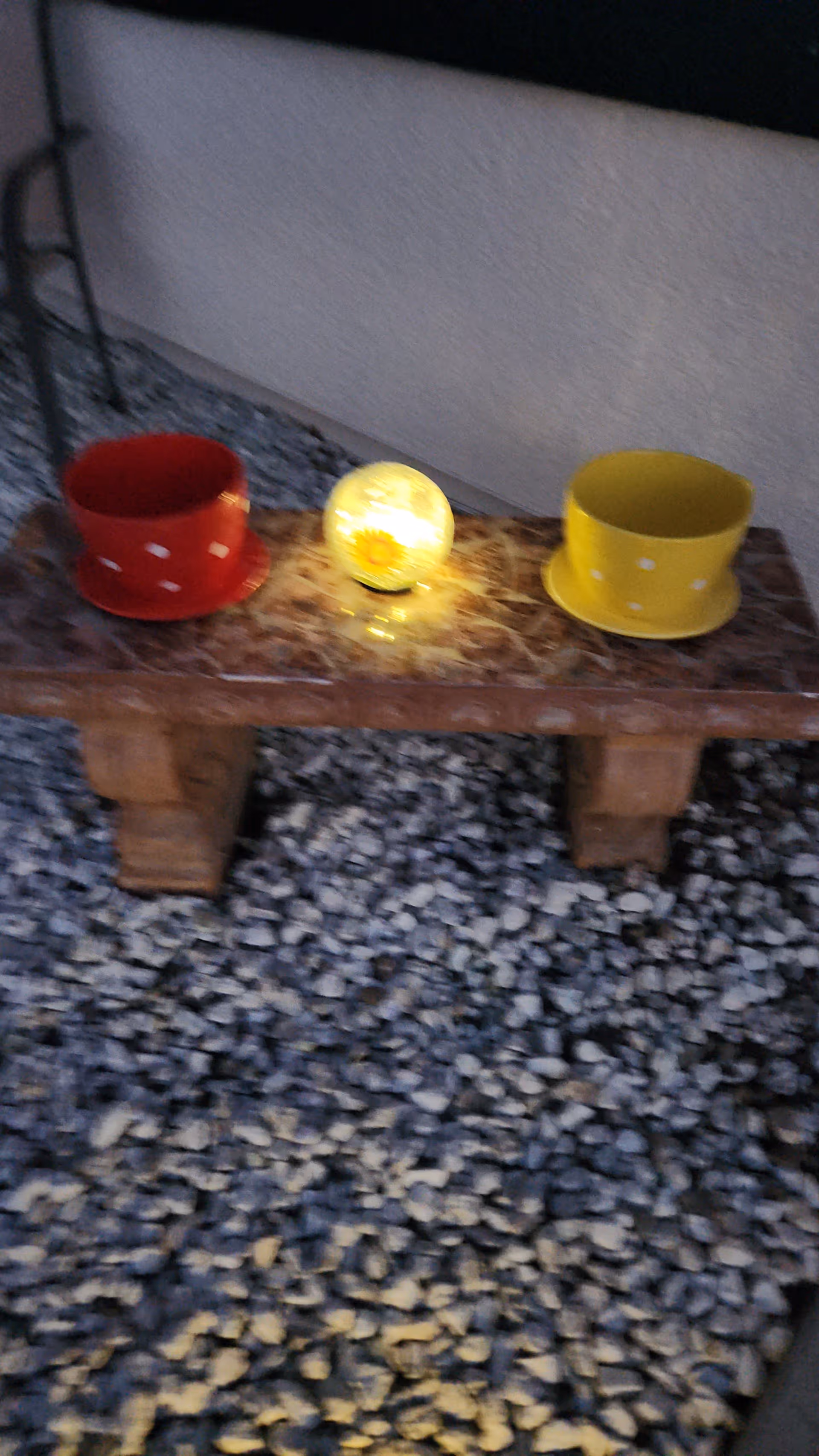 A small outdoor stone bench with a red polka-dotted cup and saucer on the left, a yellow polka-dotted cup and saucer on the right, and a glowing decorative light with a sunflower design in the center. The bench is placed on a gravel surface against a light-colored wall.