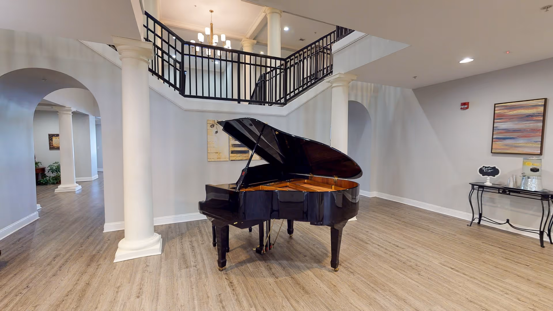 A spacious interior area featuring a black grand piano in the center, surrounded by light gray walls and wooden flooring. The room has white columns and an upper balcony with black railings. There is a small table with a water dispenser and cups on the right side, and a colorful abstract painting on the wall above it.