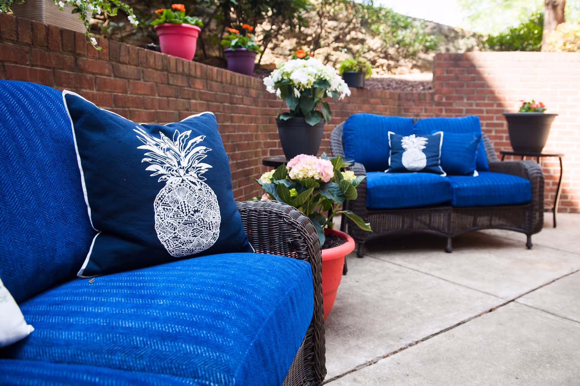 Outdoor patio area with two wicker loveseats featuring blue cushions and decorative pillows with white pineapple designs. Several potted plants with colorful flowers are placed around the seating area, and a brick wall encloses the space.