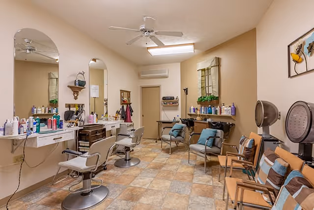 Interior view of a salon area in a senior living facility with multiple styling chairs, mirrors, shelves with hair care products, and a seating area with chairs and cushions. The room has beige walls, tiled floor, ceiling fan, and a window with a decorative valance and plants.