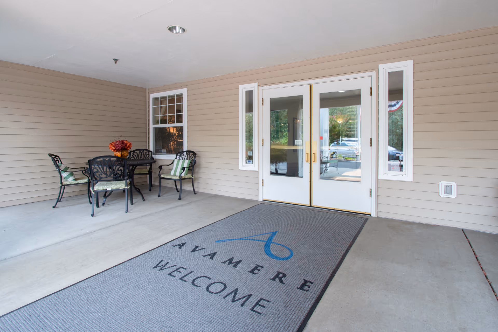 Covered entrance area with a set of black metal chairs and a table with a flower arrangement. The entrance has double white doors with glass panels and a welcome mat that reads 'AVAMERE WELCOME'.