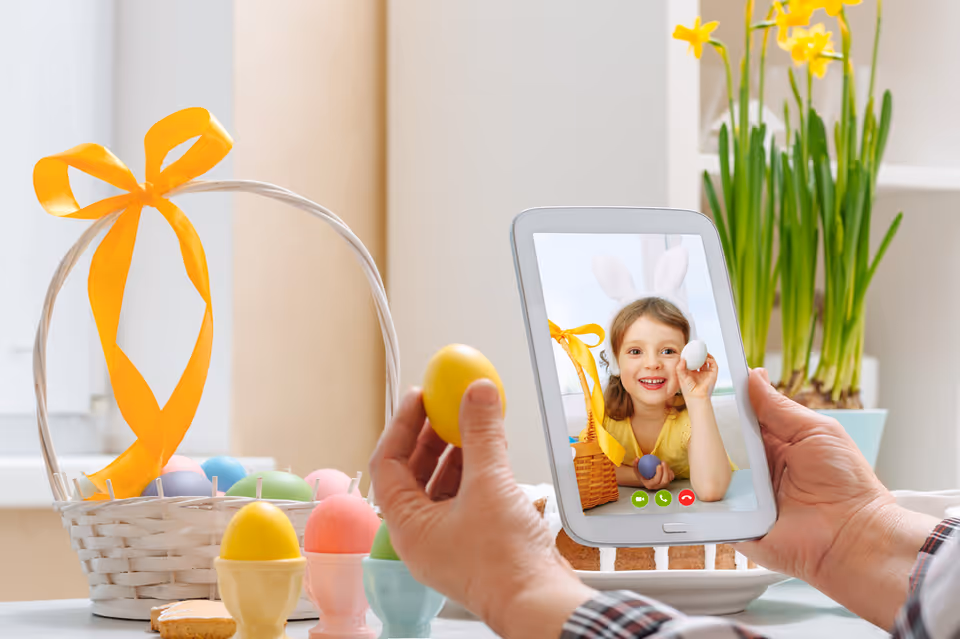 A person holding a tablet showing a video call with a smiling child wearing bunny ears and holding a white egg. In the foreground, the person holds a yellow egg. On the table, there is a white basket with a yellow ribbon filled with colorful Easter eggs, some cookies, and a vase with yellow flowers in the background.