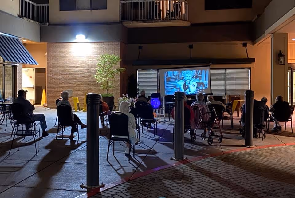 A group of elderly people sitting on chairs outdoors at night, watching a black-and-white movie projected on a screen. The setting appears to be a courtyard or patio area of a building, with some potted plants and lit windows in the background.