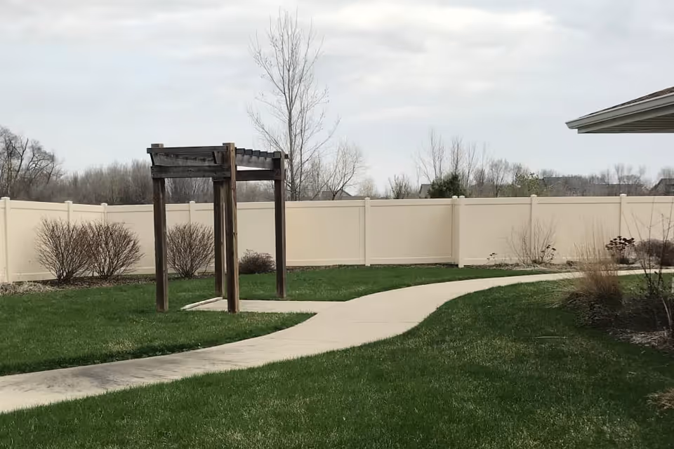 Outdoor area with a curved concrete pathway running through green grass, a wooden pergola structure, some leafless bushes, and a beige privacy fence under a cloudy sky.
