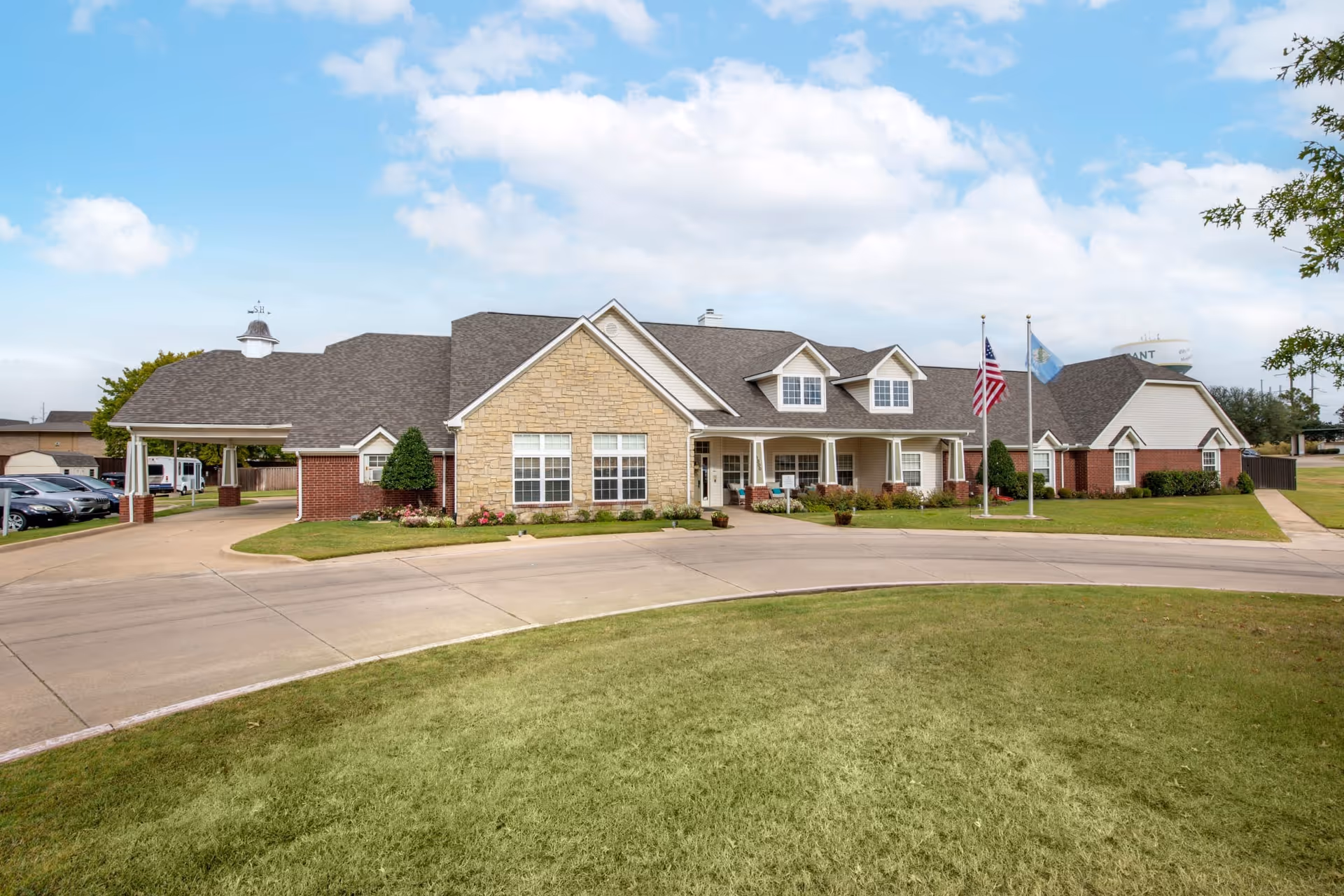 Exterior view of Oxford Springs Durant, a single-story building with a combination of brick and stone facade, multiple windows, and a covered entrance. There are two flagpoles with the American flag and another flag in front of the building. The driveway curves around a grassy area in the foreground under a partly cloudy sky.