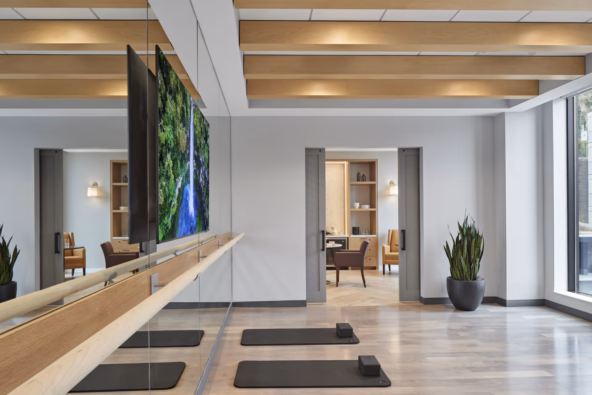 A modern exercise room with two black yoga mats and blocks on a light wood floor. A large mirror with a wooden ballet barre runs along one wall, reflecting a flat-screen TV displaying a waterfall scene. The ceiling has wooden beams, and a large window lets in natural light. Through a doorway, a cozy sitting area with chairs and shelves is visible.