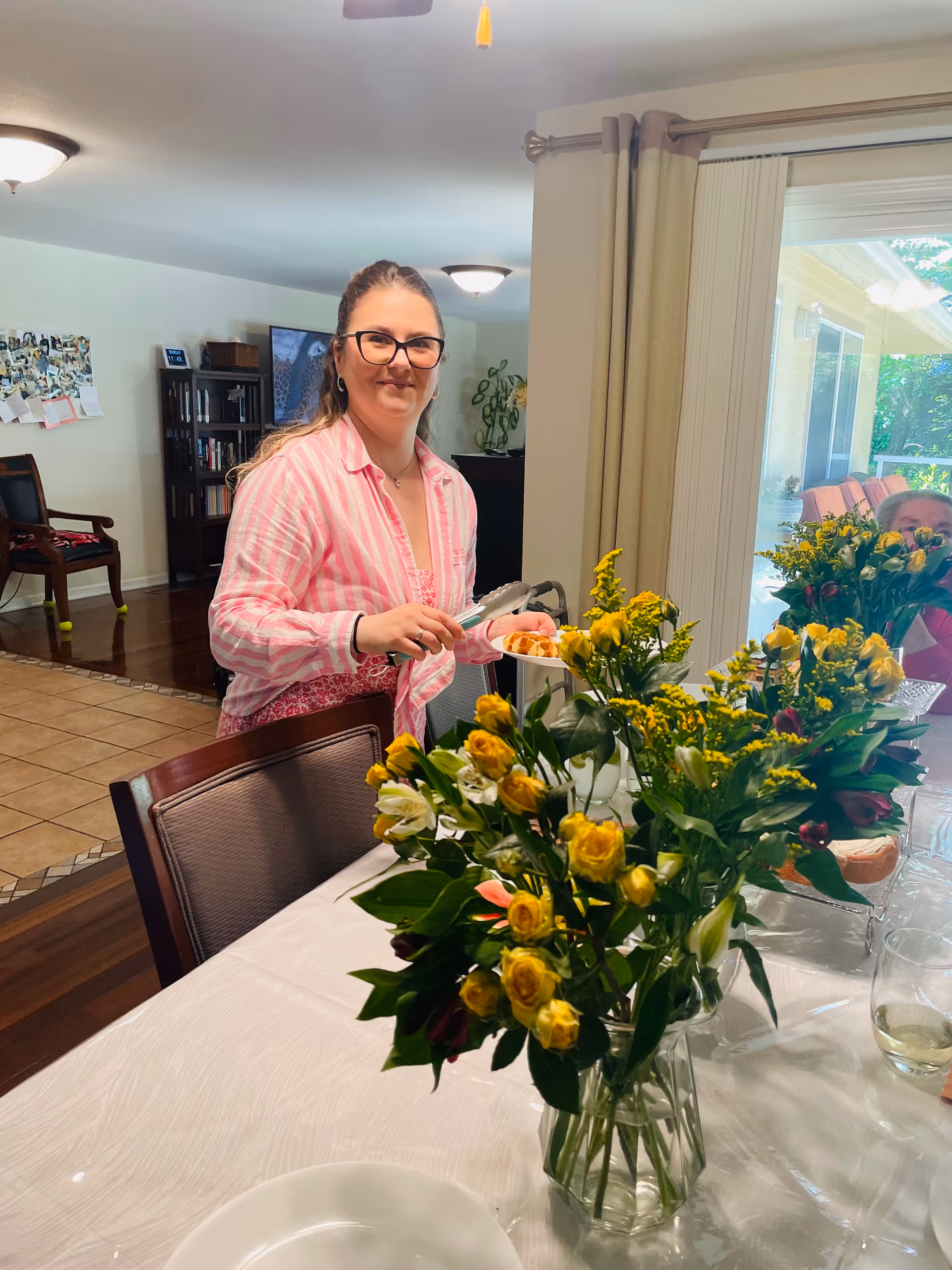 A woman wearing glasses and a pink striped shirt is standing in a dining area holding tongs and serving food onto a plate. The table in front of her is covered with a white tablecloth and decorated with several vases of yellow and white flowers. In the background, there is a living space with chairs, a bookshelf, and a sliding glass door leading to an outdoor area.