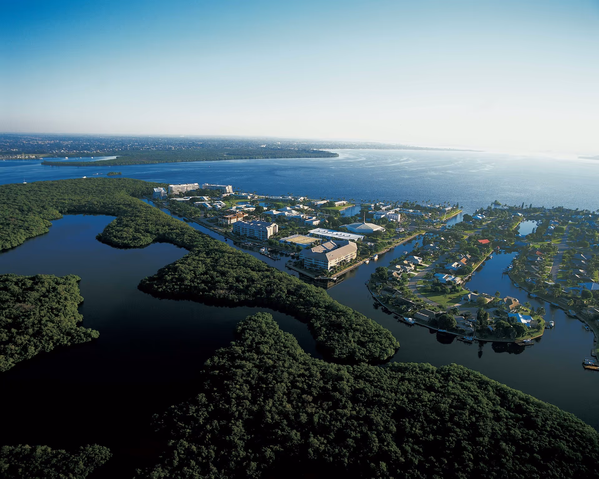 Aerial view of Shell Point Retirement Community surrounded by water and lush greenery, with residential buildings, waterways, and a large body of water extending into the horizon under a clear sky.