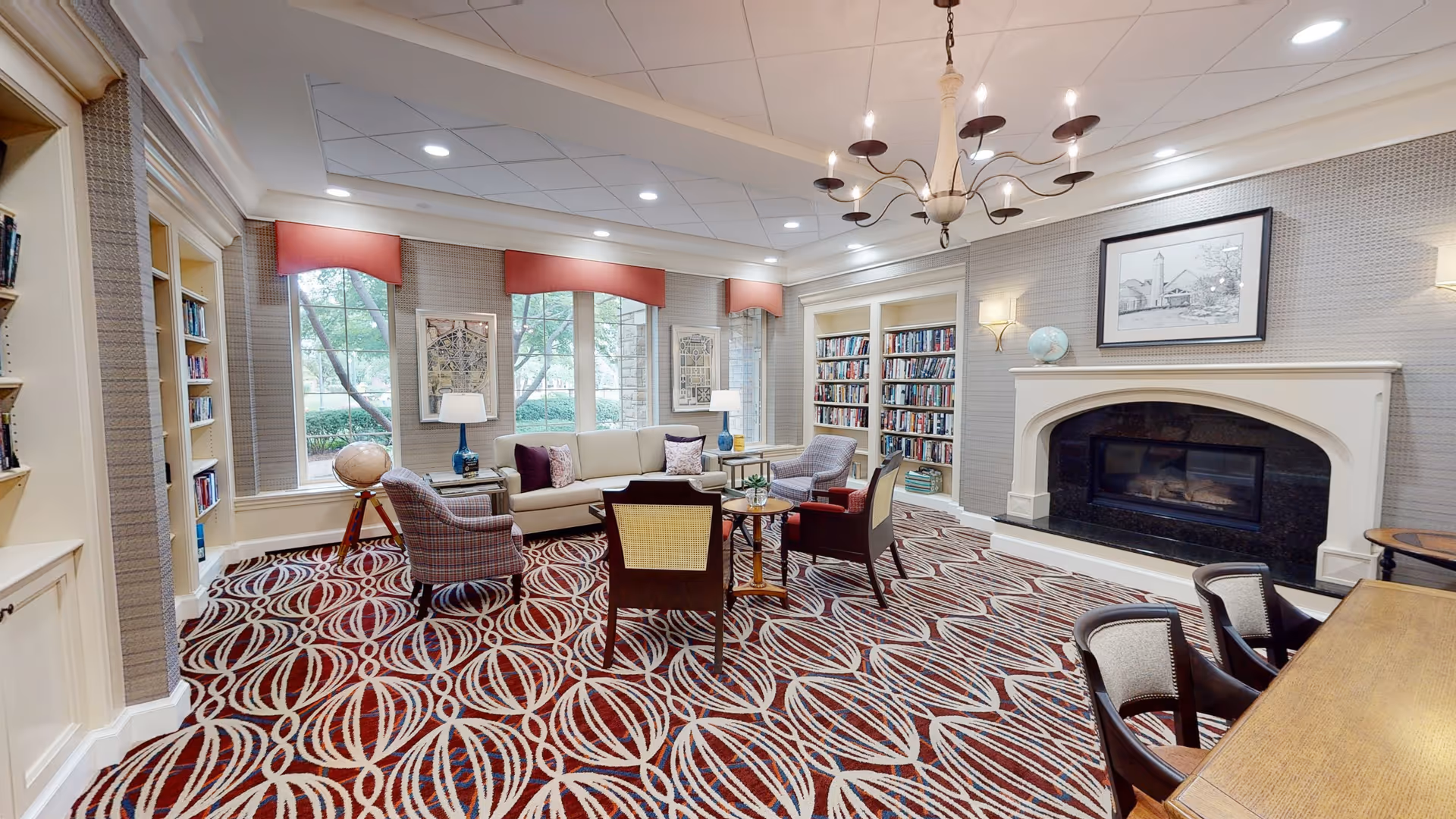 Bright communal living room with sofas and chairs arranged around a coffee table, bookshelves, a fireplace, and patterned carpet.