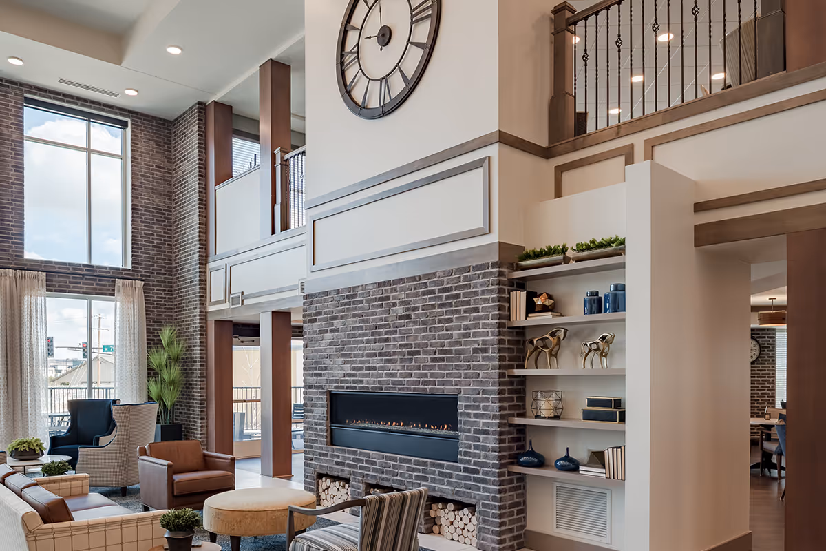 A spacious and well-lit living room area with a large brick fireplace, a big wall clock above it, and built-in shelves decorated with books, vases, and sculptures. The room features comfortable seating including leather and fabric chairs, a round ottoman, and large windows with curtains letting in natural light. There is a balcony railing visible on the upper level and a glimpse of an outdoor patio through the windows.