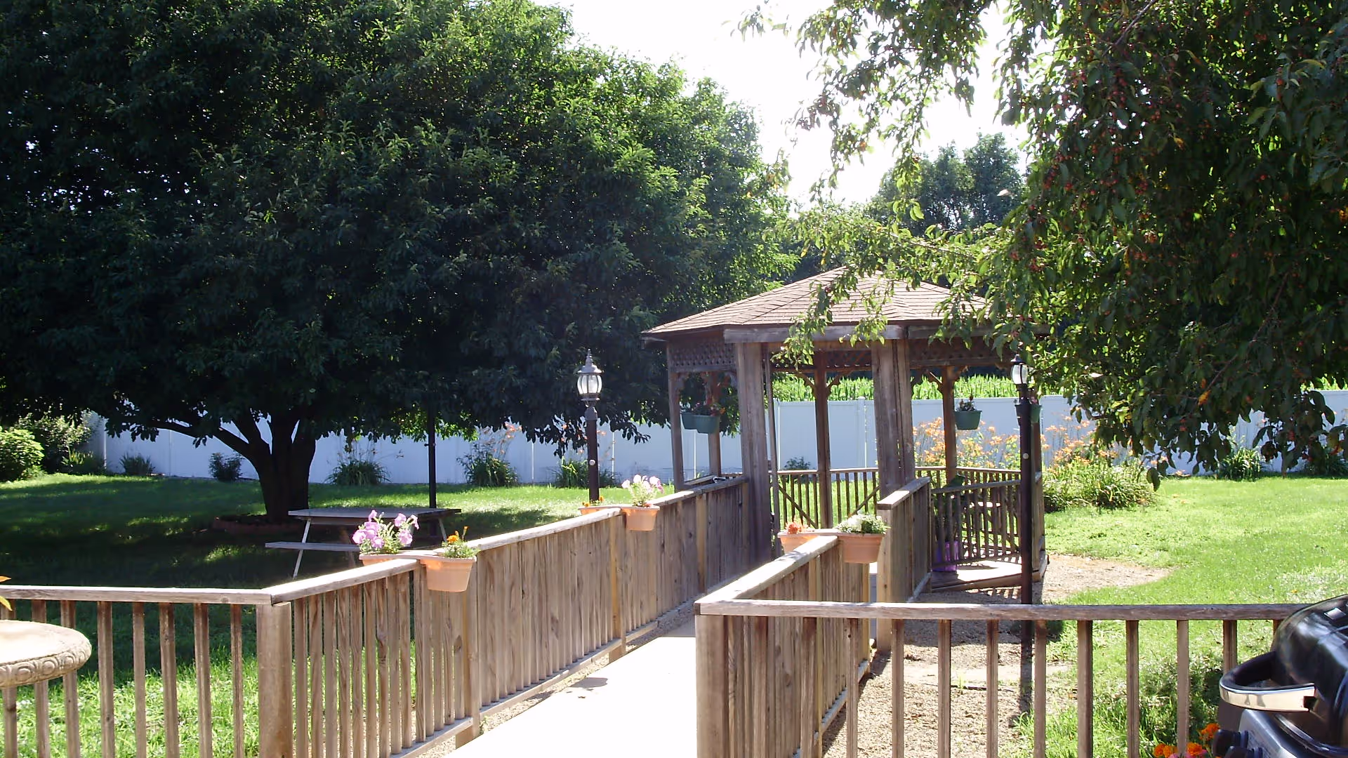 Outdoor garden area with a wooden ramp leading to a gazebo. The ramp has flower pots attached to the railing. There is a large tree providing shade, a picnic table under the tree, and a barbecue grill on the right side. The area is surrounded by green grass and a white fence in the background.