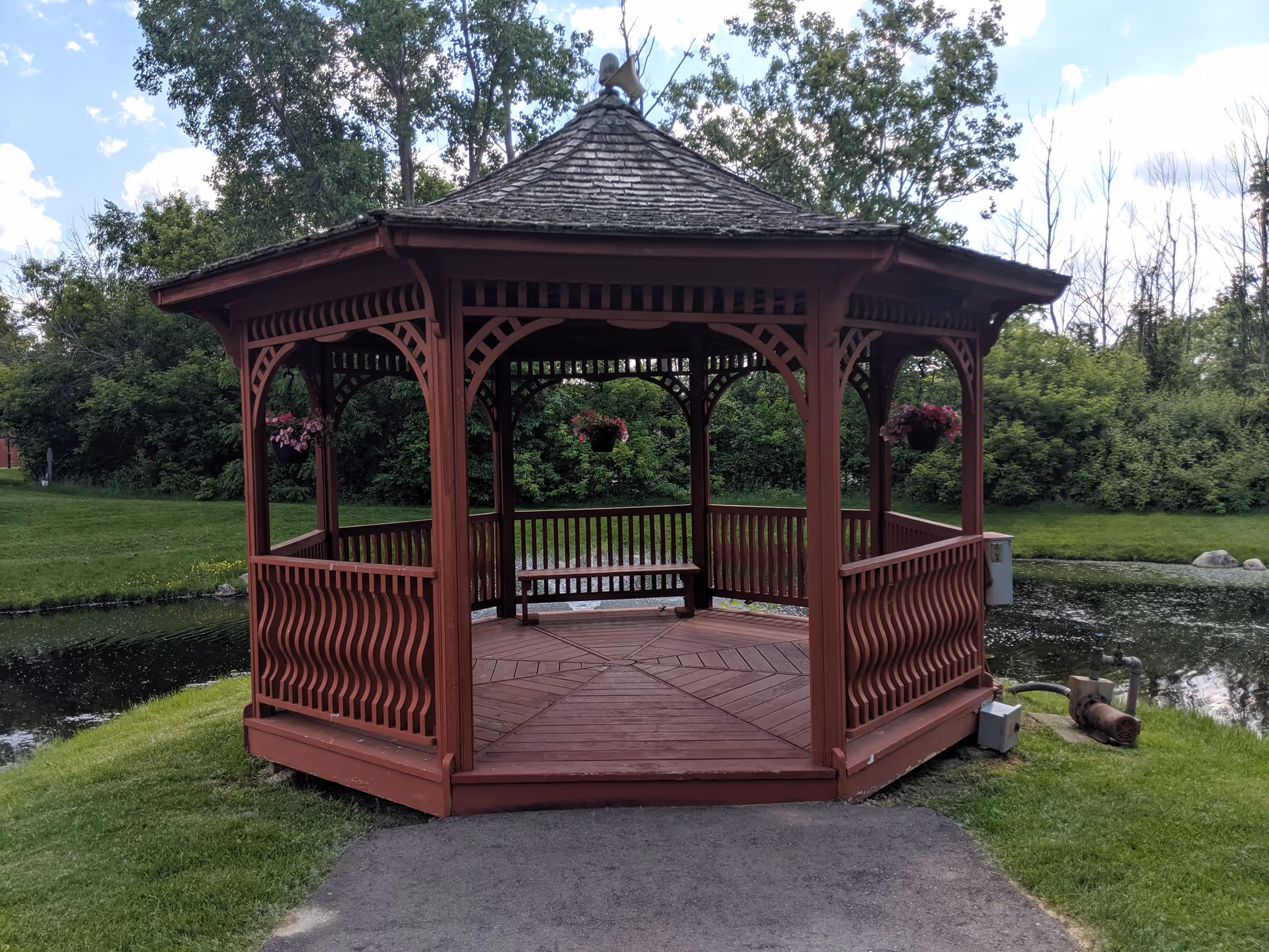 A red wooden gazebo with a shingled roof situated on a grassy area next to a small pond, surrounded by trees and bushes under a partly cloudy sky.