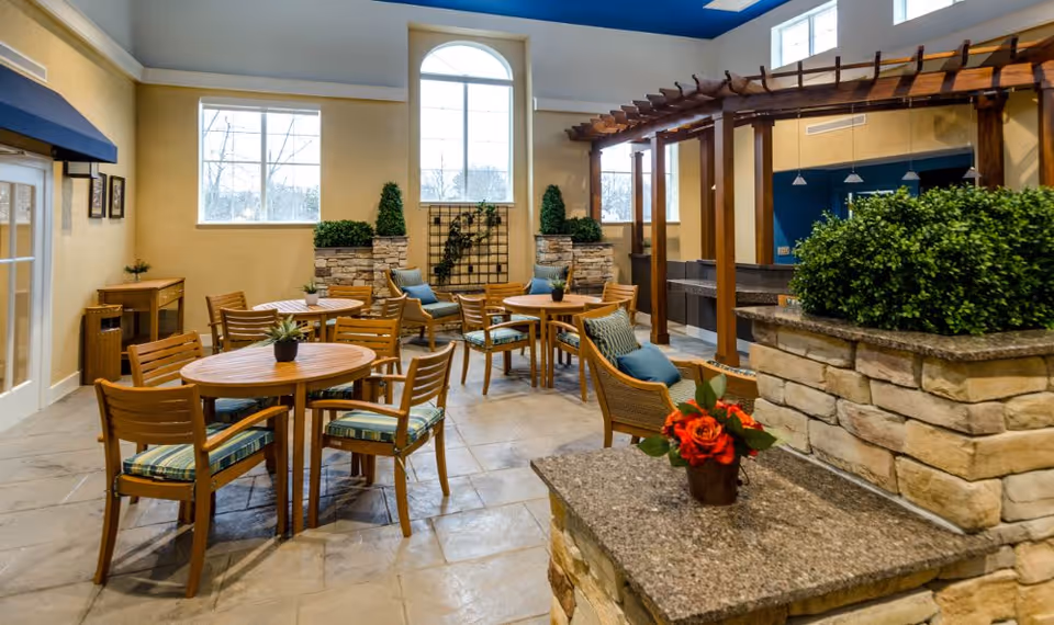 Indoor seating area with round wooden tables and chairs with striped cushions, stone planters with green plants, a wooden pergola structure, and large windows letting in natural light.