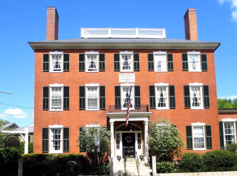 Front exterior view of a large, three-story red brick building with white trim and black shutters. The building has two prominent chimneys, a central entrance with a small portico, and an American flag hanging above the door. There are bushes and greenery in front of the building, and a sign on the left side that reads John Bertram House.