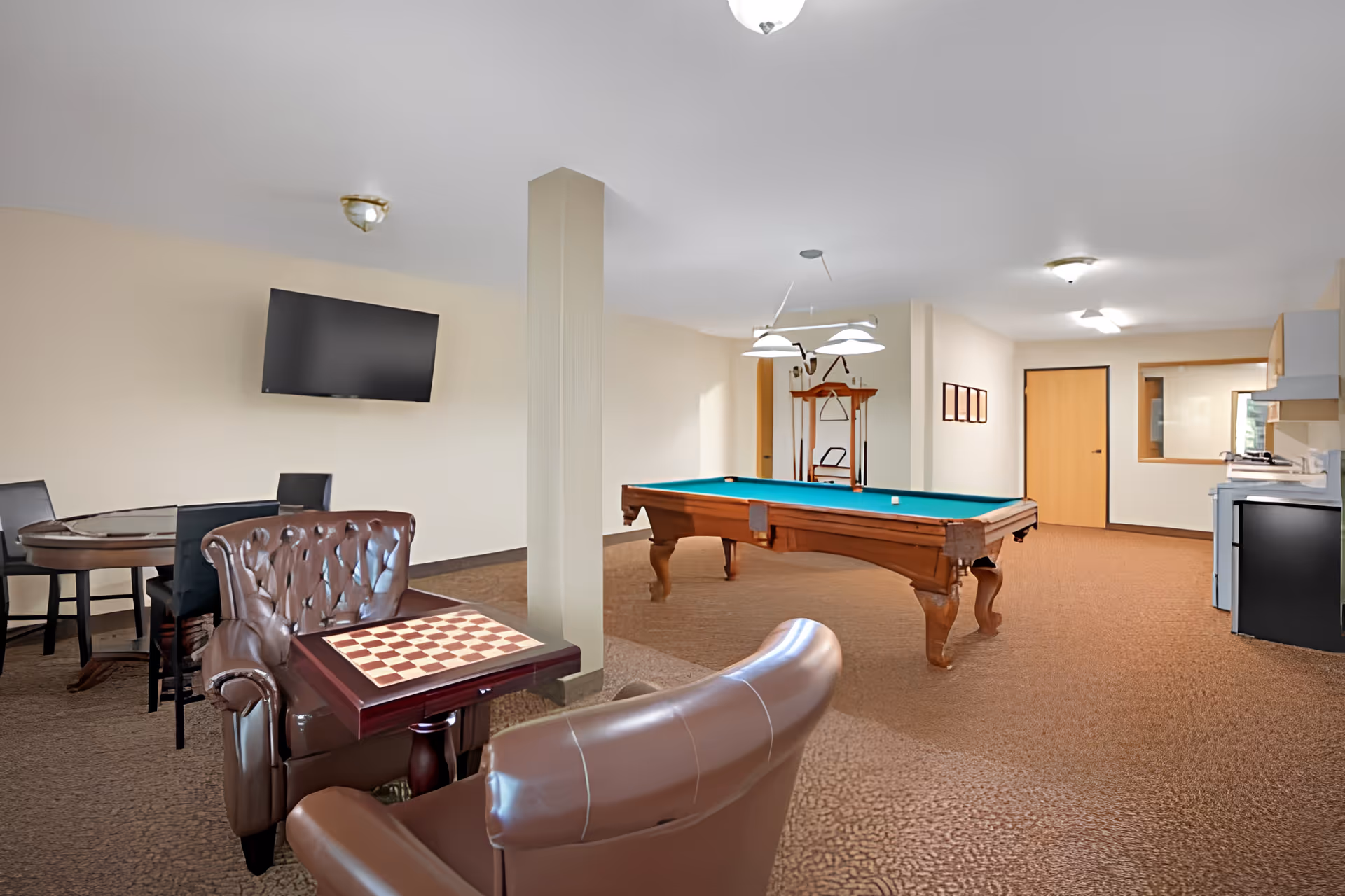 Recreation room featuring a pool table, leather armchairs with a chessboard, a card table, wall-mounted TV, and a small kitchenette.