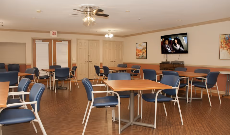 A spacious common area with multiple wooden tables and blue cushioned chairs arranged neatly on a wooden floor. The room has beige walls with two framed paintings and a flat-screen TV mounted on the wall showing a scene from a movie or TV show. There are double doors with white blinds and an exit sign above them. The ceiling has two light fixtures and a ceiling fan with lights.