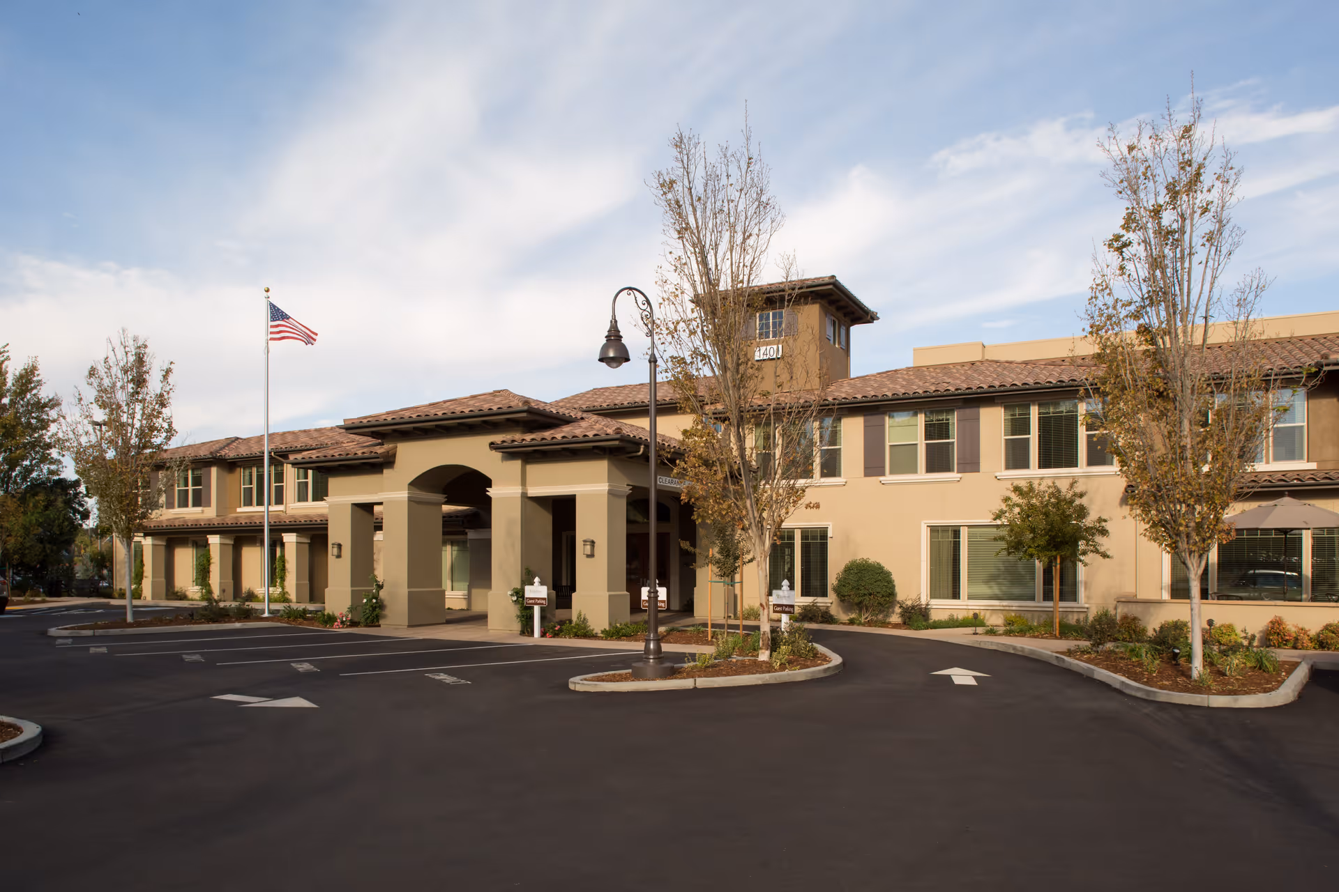 Exterior view of Oakmont of Concord senior living facility showing a two-story building with beige walls and brown roof tiles, surrounded by small trees and landscaping. There is a flagpole with an American flag and a paved driveway with directional arrows leading to the entrance.