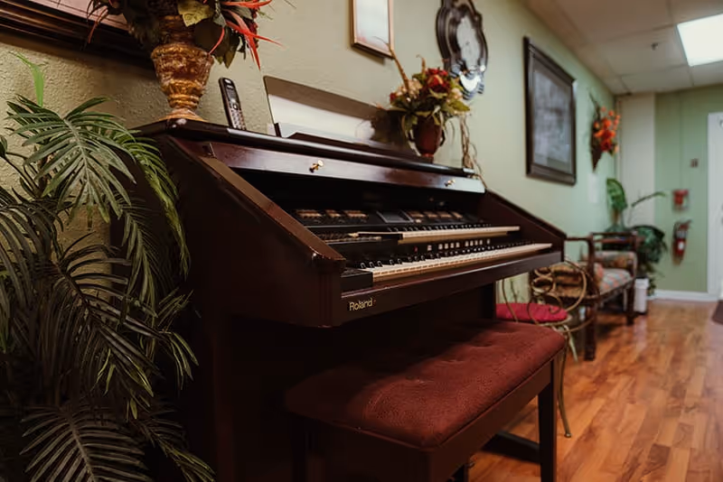 A wooden electric organ with a bench sits in a decorated hallway/common area with plants, seating, and framed wall art.