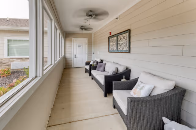 A covered outdoor patio area with wicker furniture including a cushioned armchair and a sofa. The patio has beige walls with horizontal paneling, two ceiling fans, and large windows on one side overlooking a garden area.