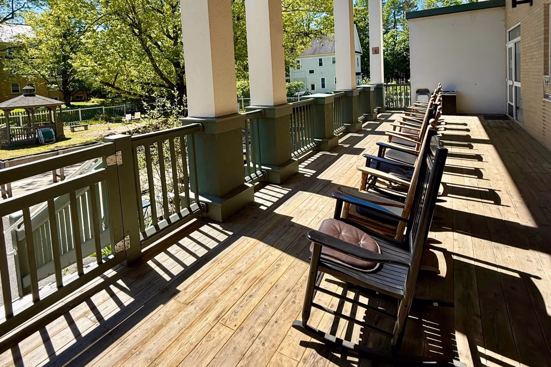 An outdoor wooden balcony with a row of rocking chairs along the railing overlooking a yard with trees.