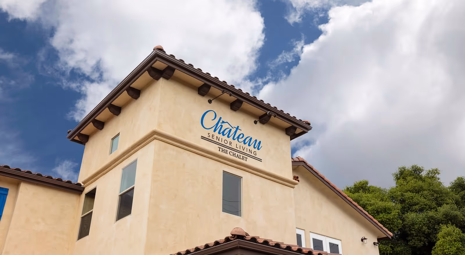 Exterior view of a beige stucco building with a tiled roof under a partly cloudy sky. The building has a sign that reads 'Chateau Senior Living The Chalet'.