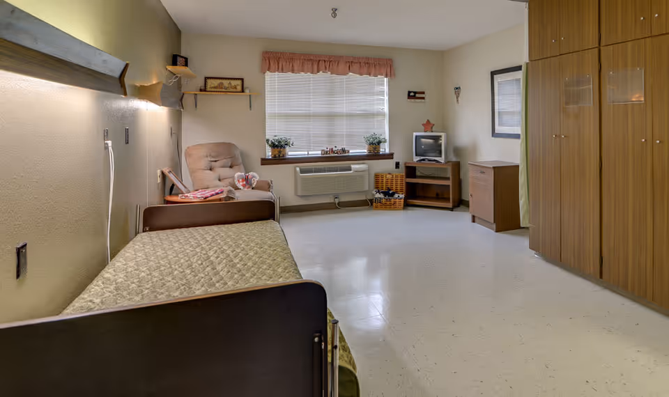 A simple, clean nursing facility room with a single bed covered in a green quilt, a beige recliner chair, a small wooden TV stand with an old-style television, a window with blinds and a valance, and wooden wardrobes along one wall. The floor is light-colored and the walls are painted beige.