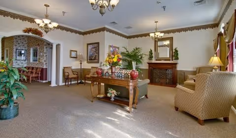 A spacious and well-lit living room area in a senior living facility with beige carpet, comfortable upholstered chairs, a wooden table with floral arrangements, a fireplace with a mirror above it, and decorative plants. The room has multiple light fixtures and large windows with curtains allowing natural light to enter.