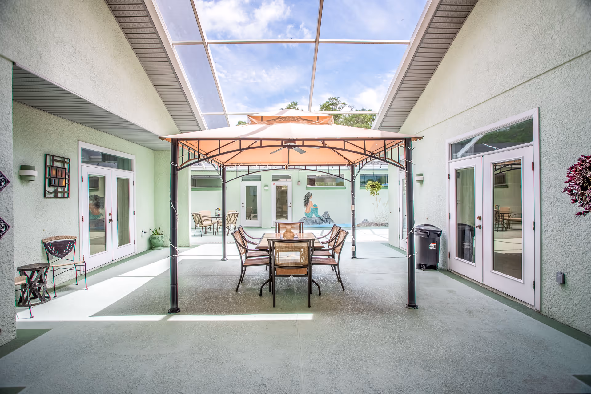A covered outdoor patio area with a table and six chairs under a beige canopy. The patio is enclosed by light green walls with multiple glass doors and windows. There is a mural of a mermaid on the far wall and some potted plants and decorative items along the walls. The ceiling above the patio is a transparent screened enclosure allowing natural light to come through.