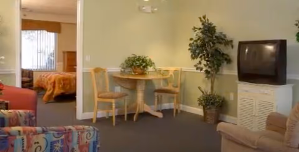Cozy assisted-living living room with chairs, a small round table and plants, a TV on a cabinet, and a bedroom visible through an open doorway.