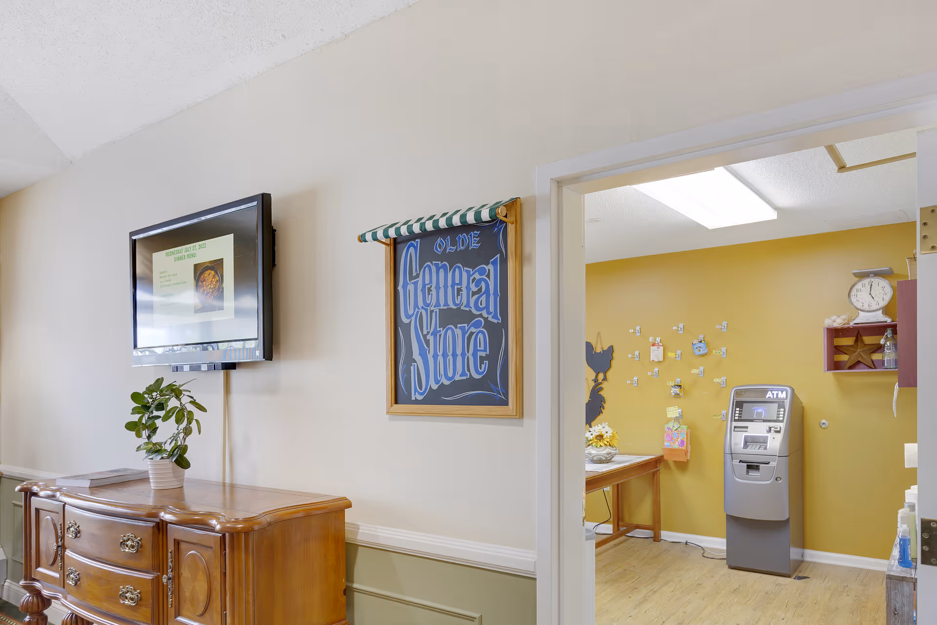 Interior view of a senior living facility hallway with a wooden sideboard holding a potted plant and papers. A flat-screen TV is mounted on the wall above the sideboard. Next to the TV is a framed sign that reads 'Olde General Store'. Through an open doorway, a room with yellow walls is visible, containing an ATM machine, a wooden table with decorations, and wall-mounted items including a scale and a star decoration.