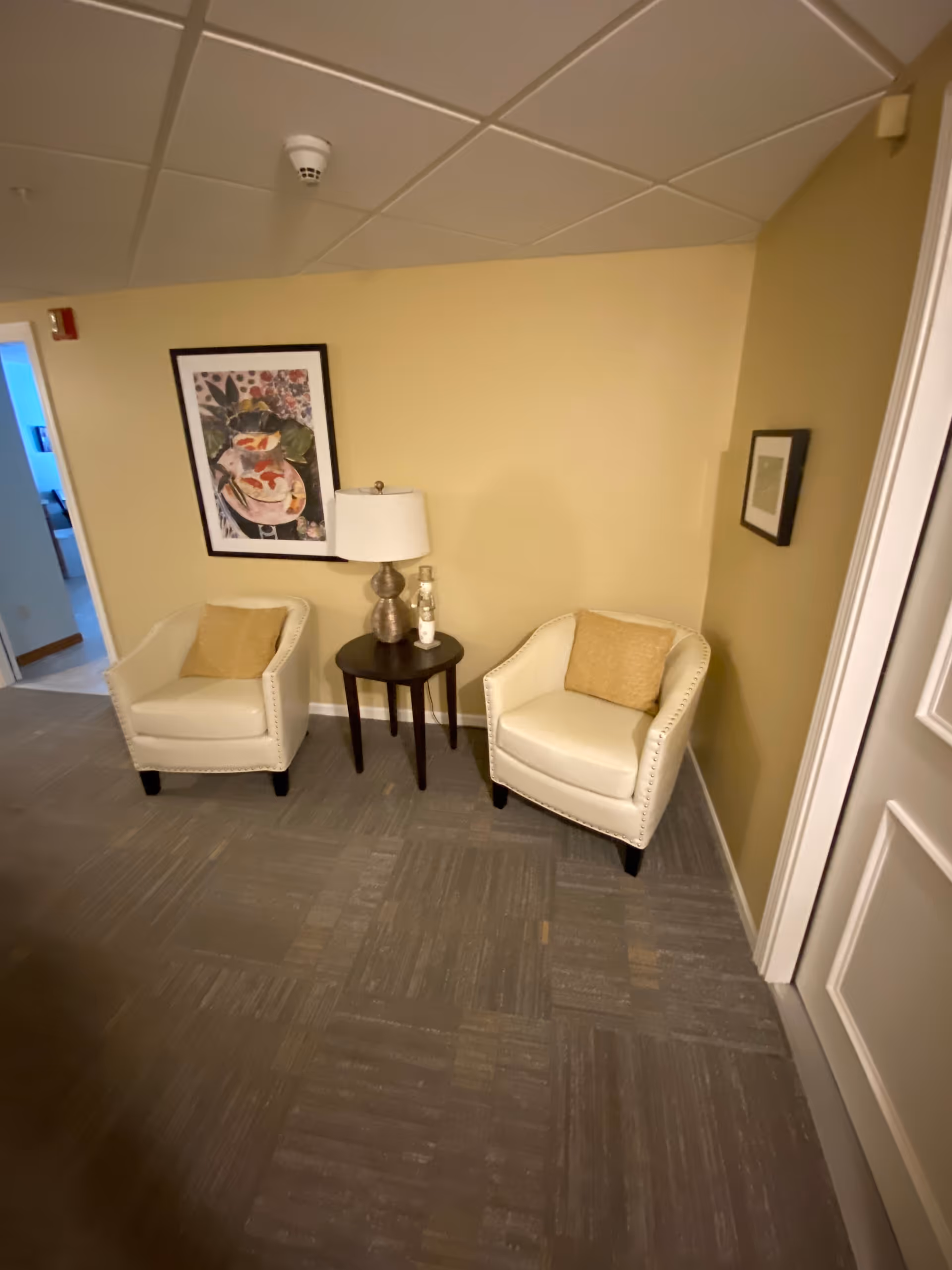 A small seating area with two white armchairs, each with a beige cushion, positioned on either side of a dark wooden side table holding a lamp and a small decorative item. The walls are painted light yellow and decorated with two framed pictures. The floor is covered with gray carpet tiles.