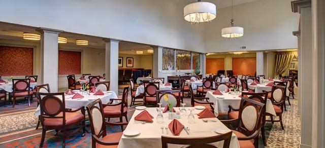 Spacious dining room with multiple tables set with white tablecloths, folded red napkins, wooden chairs, and hanging chandeliers.