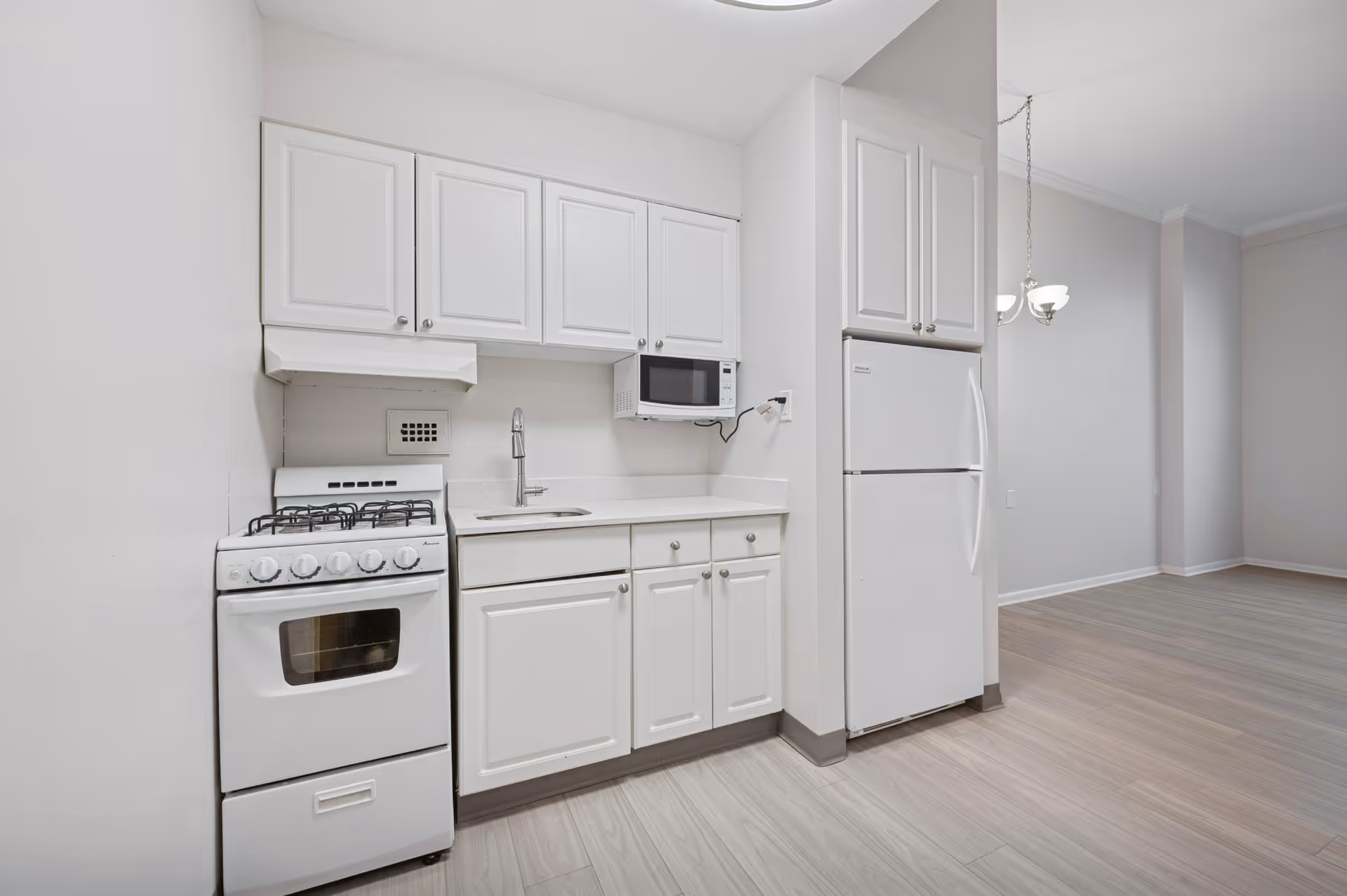 A clean, modern kitchen with white cabinetry, a white gas stove, a white refrigerator, a microwave mounted above the counter, and a stainless steel sink. The kitchen opens into a dining area with a chandelier and light wood flooring throughout.