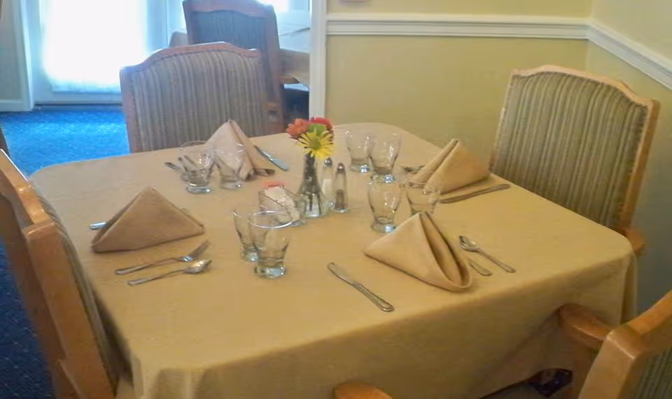 A dining table set for four with beige tablecloth, folded beige napkins, drinking glasses, silverware, and a small vase with flowers in the center. The table is surrounded by four upholstered chairs with wooden frames. The background shows part of the room with a blue carpet and light-colored walls.