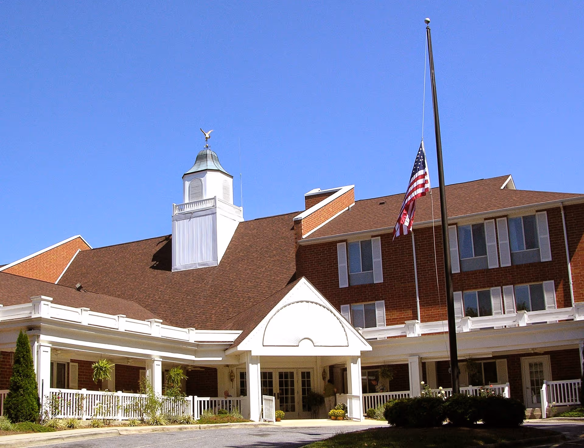 Front exterior of a brick senior living facility showing the covered entrance with white columns, a roof cupola, and an American flag at half-staff.