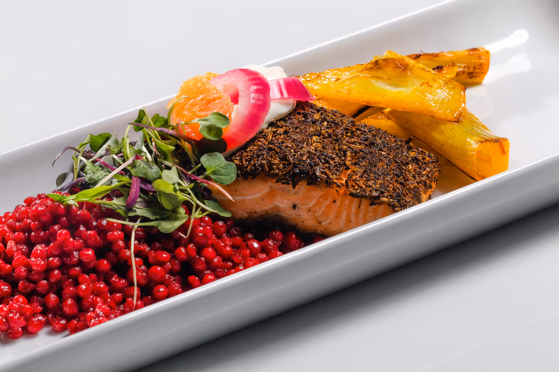 A plated gourmet meal featuring a herb-crusted salmon fillet, roasted yellow vegetables, a garnish of microgreens, pickled red onions, a dollop of cream, and a side of red berries or small red fruit on a white rectangular plate.