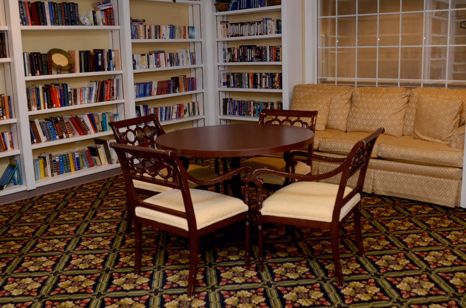 A cozy interior room featuring a round wooden table surrounded by four cushioned wooden chairs. Behind the table, there is a beige patterned sofa with multiple cushions. The walls are lined with white bookshelves filled with various books. The floor is covered with a patterned carpet in dark colors with floral designs.