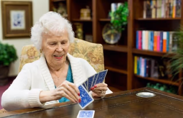 An elderly woman with white hair sitting at a wooden table, smiling and holding playing cards in her hands. Behind her is a bookshelf filled with books and decorative items, and a framed picture on the wall.