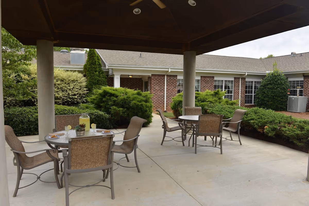 Outdoor covered patio area with two round tables, each surrounded by four chairs. One table has a pitcher of lemonade and glasses with lemon slices. The patio is surrounded by green bushes and trees, with a brick building in the background.