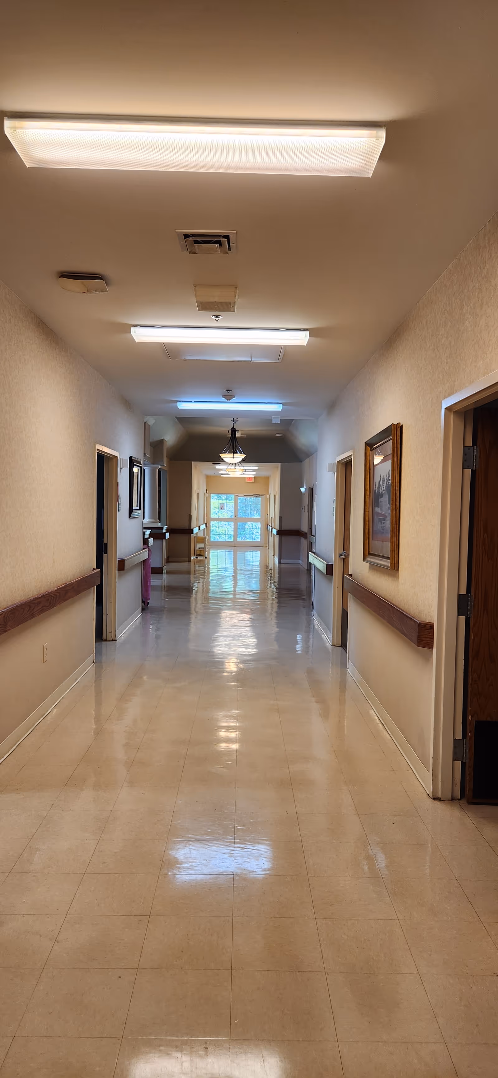A long, clean hallway in a healthcare facility with beige walls, handrails on both sides, several doorways, framed pictures on the walls, and fluorescent ceiling lights. At the end of the hallway, there is a window letting in natural light.