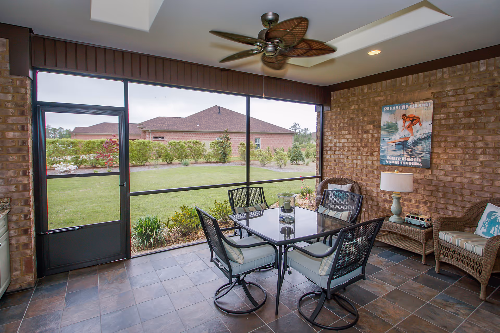 A screened-in patio area with a glass-top table surrounded by four cushioned metal chairs. There are two wicker armchairs with cushions and a wicker side table with a lamp and decorative items. A ceiling fan is mounted above, and a vintage-style surfing poster is on the brick wall. Outside the screened area, there is a well-maintained lawn and shrubs with neighboring buildings in the background.