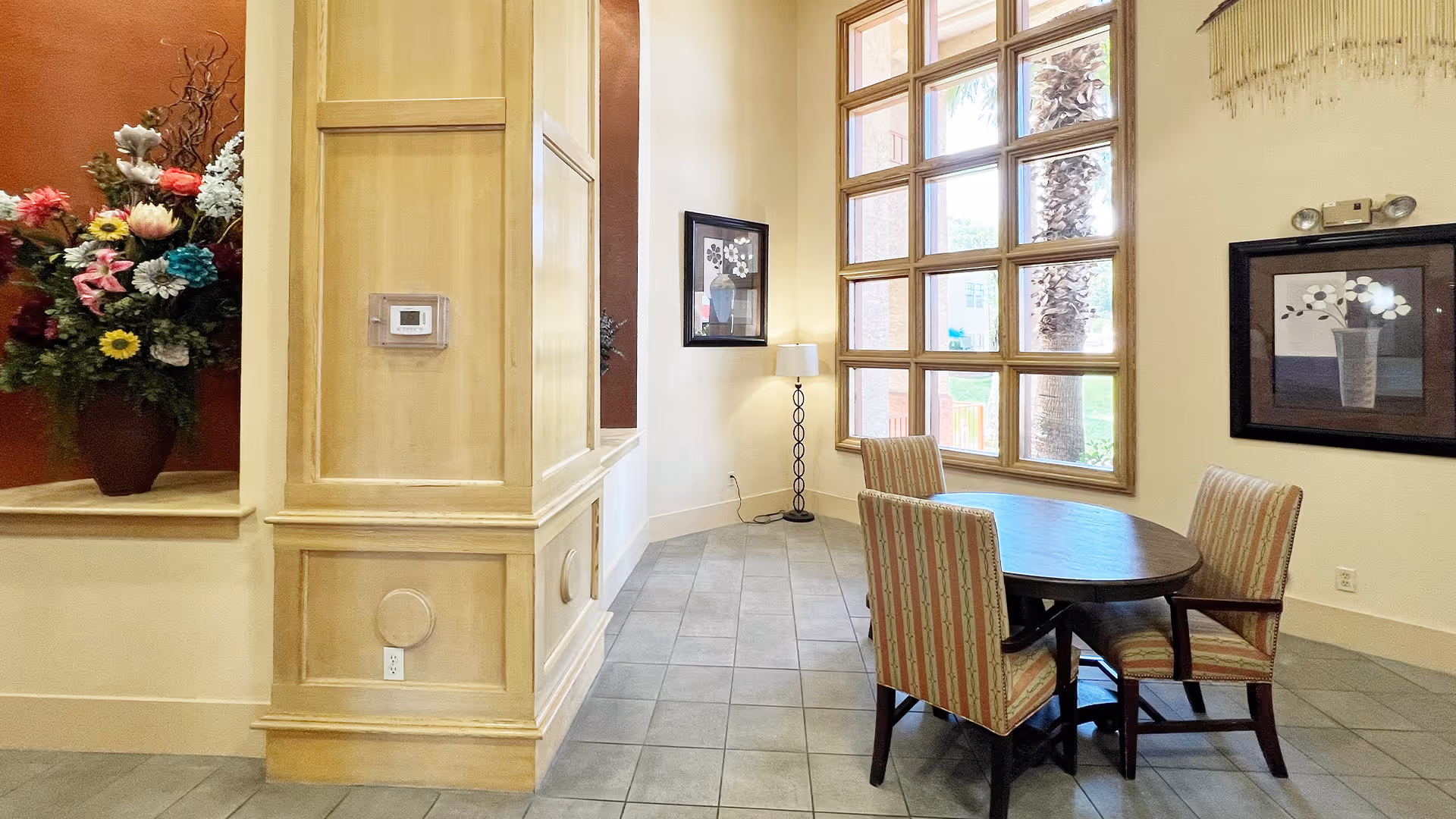 A bright interior corner of a senior living facility with a round wooden table surrounded by four striped upholstered chairs. Large windows with wooden frames let in natural light and show palm trees outside. The walls are decorated with framed floral artwork, and a floor lamp stands in the corner. A large floral arrangement is placed on a ledge against a warm-toned wall.
