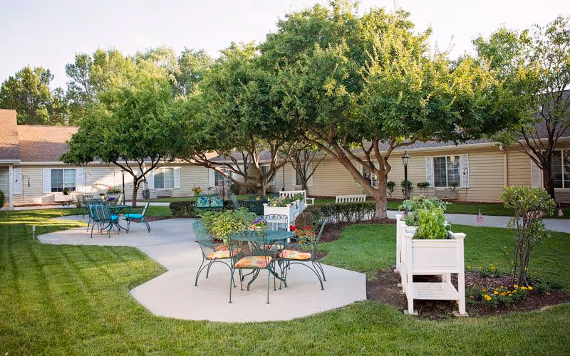 Outdoor patio area at Bickford of Lincoln with green metal tables and chairs, some with floral cushions, surrounded by well-maintained grass, trees, and planter boxes with flowers and greenery. Single-story beige buildings with windows and white shutters are visible in the background.