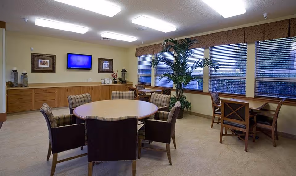 A well-lit common room with a round table surrounded by six cushioned chairs in the center. Along the wall is a long wooden cabinet with decorative items and a flat-screen TV mounted above it. There are large windows with blinds on the right side, letting in natural light, and a tall potted plant near the windows. Additional smaller tables with chairs are placed near the windows.