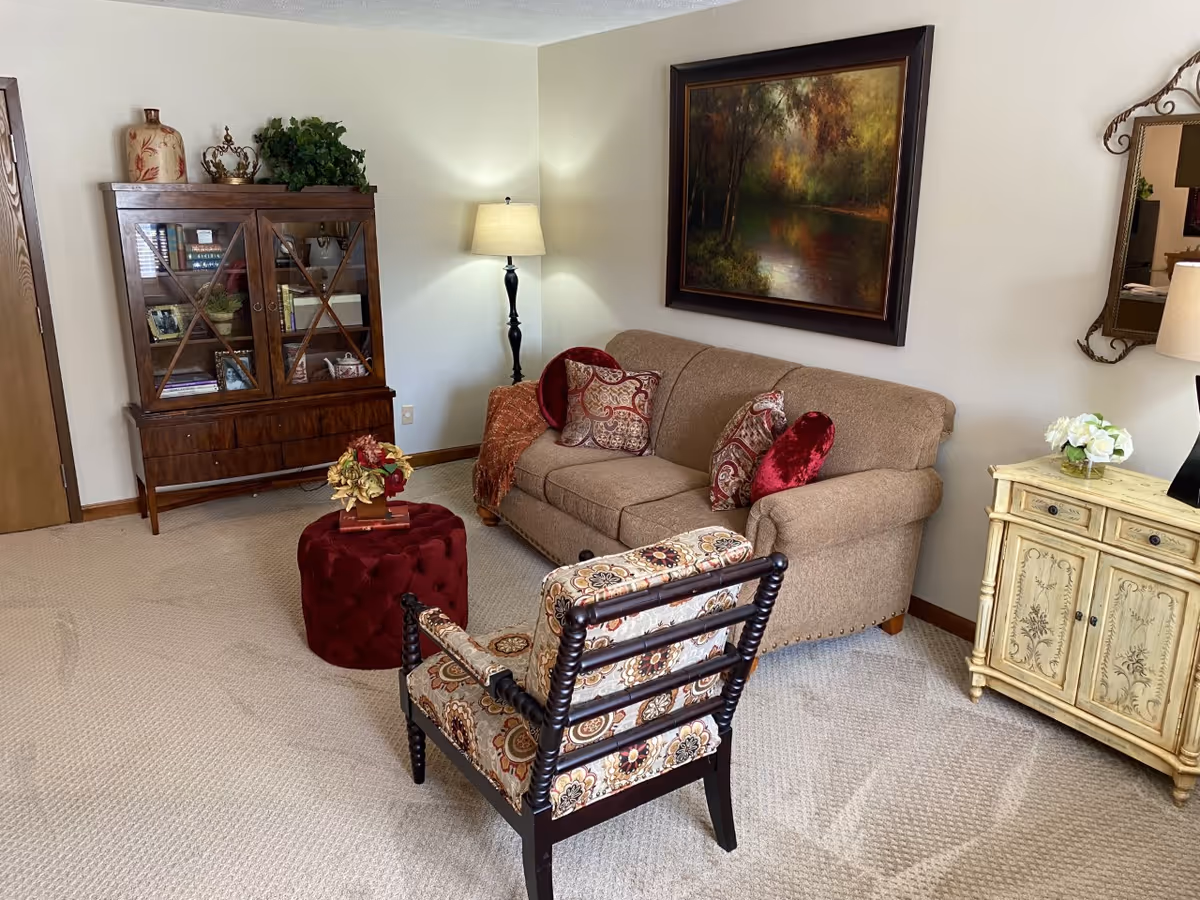 A cozy living room with a beige sofa adorned with patterned and red velvet pillows, a floral patterned armchair, a red tufted ottoman with a flower arrangement on top, a wooden cabinet with glass doors displaying books and decorative items, a floor lamp, a framed landscape painting on the wall, and a cream-colored side cabinet with floral designs and a small flower vase.