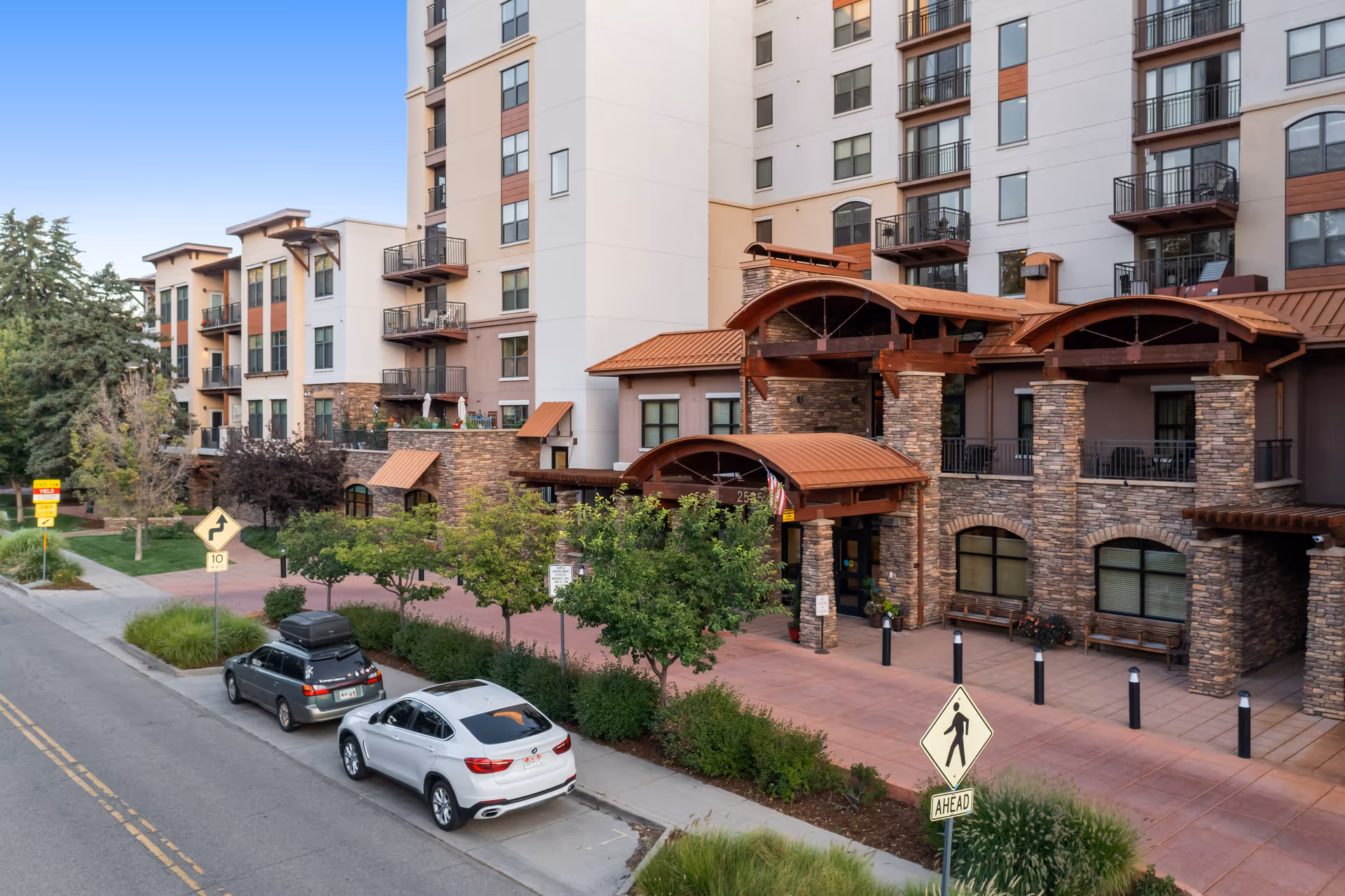 Exterior view of The Pearl at Boulder Creek senior living facility showing a multi-story building with balconies, stone and stucco facade, a covered entrance with wooden beams, parked cars along the street, and surrounding greenery under a clear blue sky.