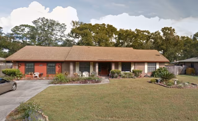 Single-story residential building with a brown shingle roof, red and beige exterior walls, several windows, and a front porch with white chairs. The building is surrounded by a well-maintained lawn, shrubs, and trees in the background. A driveway with a parked car is visible on the left side.