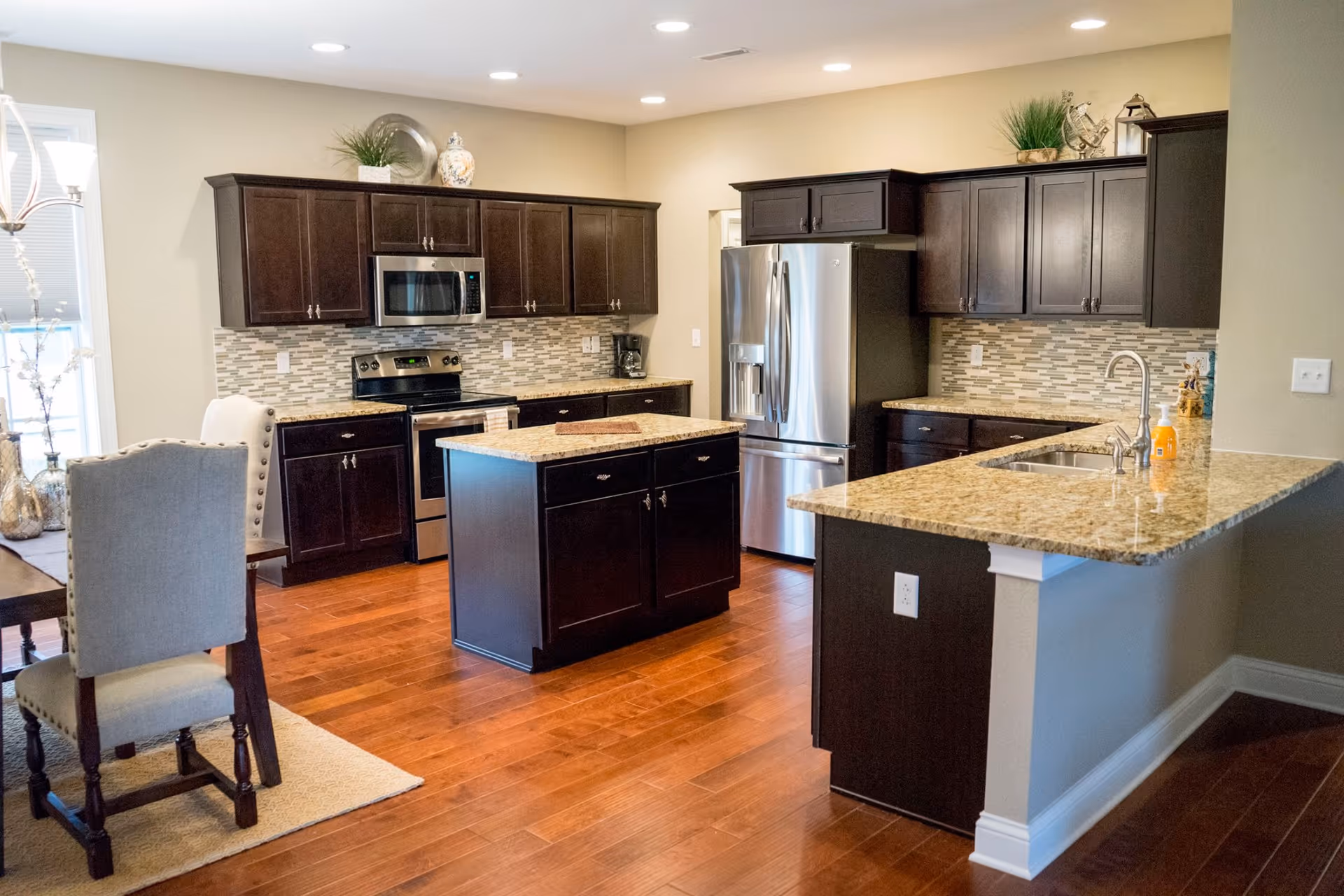Open modern kitchen with dark wood cabinets, a center island, granite countertops, stainless steel appliances, and adjacent dining chairs.