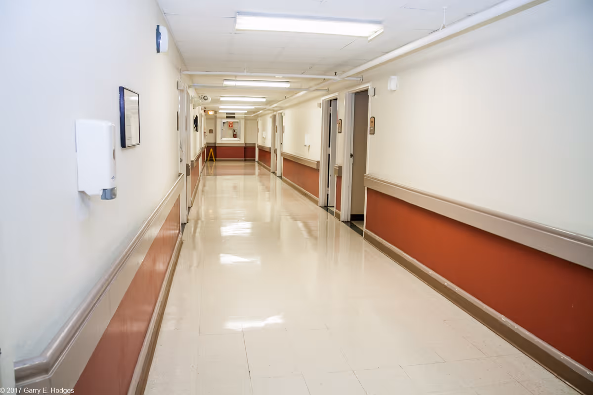 Long, clean interior corridor in a nursing facility with handrails and several open doorways.