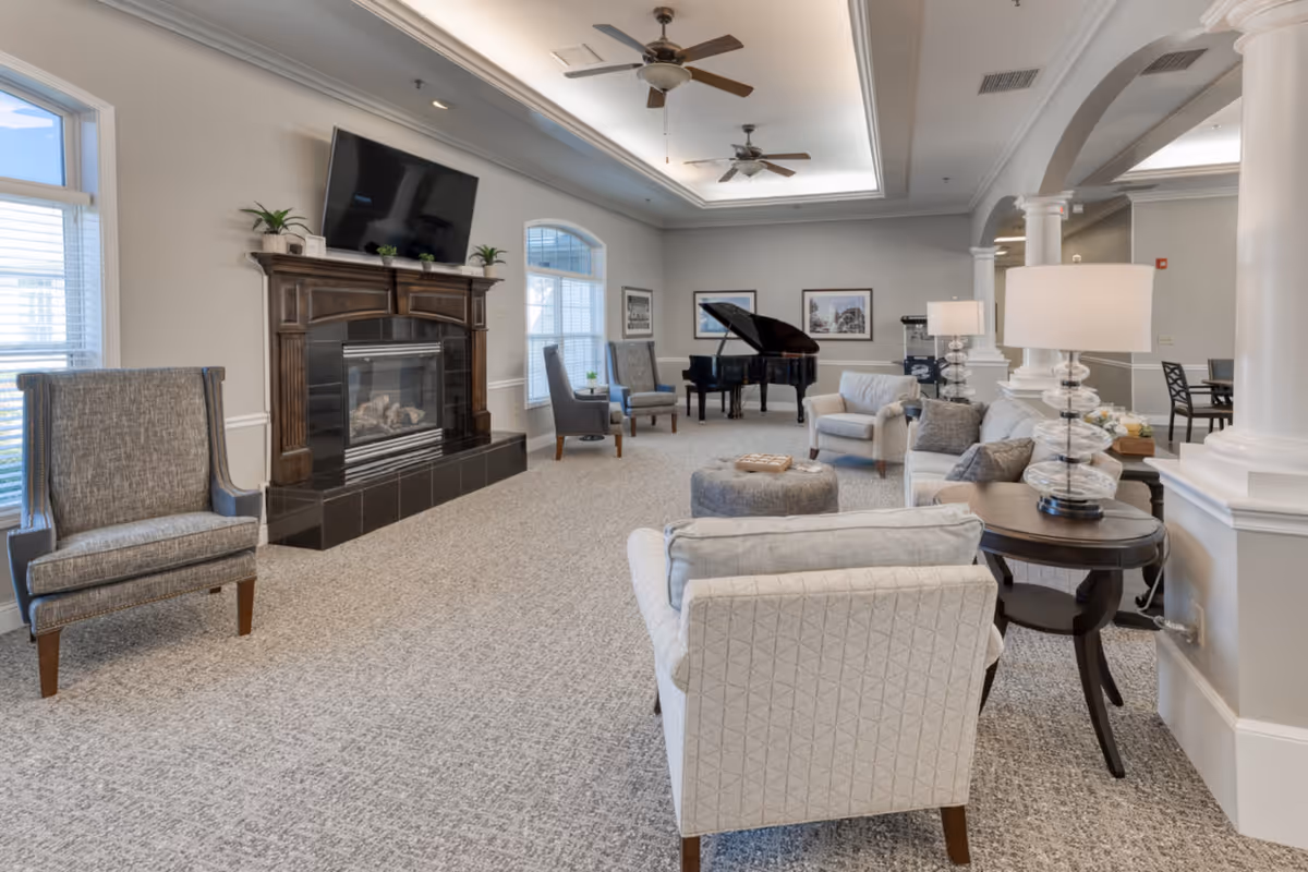 A spacious and well-lit living room area in a senior living facility featuring a fireplace with a mounted flat-screen TV above it, several upholstered armchairs and sofas arranged around a central ottoman, two table lamps on side tables, large windows letting in natural light, and a grand piano in the background near framed artwork on the walls.